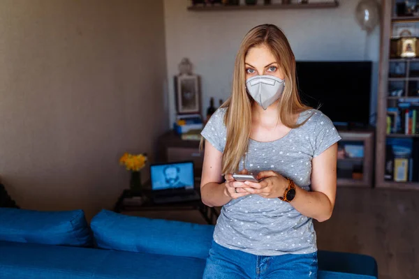 Beautiful girl reading news on the phone about the coronavirus epidemic and calling her friends. She remains isolated at home alone and wearing protective respirator while working remotely.