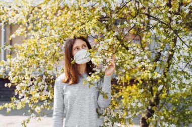 İlkbaharda çiçek açan ağacın yanında koruyucu maske takmış üzgün genç bir kadının yan görüntüsü. Bahar alerjisi. Coronavirus koruması.