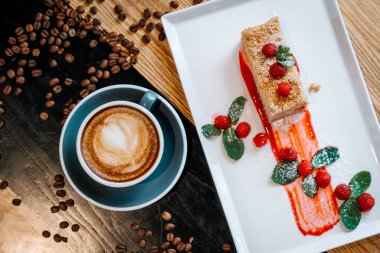Gri fincanda kapuçino sanatı, bir parça Napolyon pastası, ahududu ve nane yaprağı ile birlikte restorandaki ahşap masada kahve çekirdekleri arasında. Sabah kahvaltısı için kahve aşığı konsepti