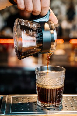 Dikey görüntü. Alternatif Kahve Hazırlama Yöntemi. Kahvecinin ellerine yakın çekim, çaydanlıktan siyah filtre kahveyi bir kafedeki barın tezgahına şeffaf bir bardağa döküyor..