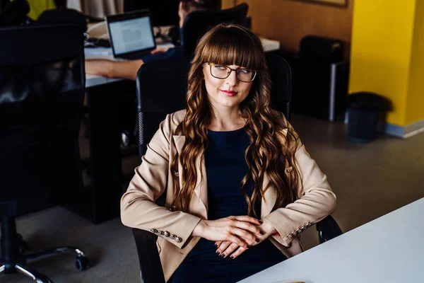 Confident, clever, business lady working with papers in office room in a coworking space. Sitting at the table, dressed in a dark blue dress and a brown jacket. She wears glasses and looks very smart