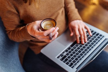 Ofiste dizüstü bilgisayar ve bir fincan aromatik espresso ve kapuçinoyla çalışan zeki bir kız. Dizüstü bilgisayarını kucağında tutuyor ve güzel bir kahve aromasının tadını çıkarıyor.