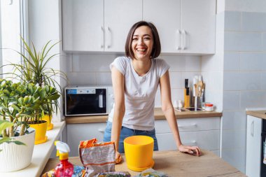 Güzel, çekici bir kız, beyaz tişört ve kot pantolon giyiyor, parlak, beyaz bir mutfakta duruyor. Masaya yaslanıp gülüyor. Masadaki çiçekleri yeniden dikmek için malzemeler var. Hazırlanıyor.