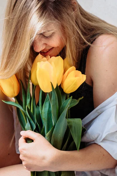 Beautiful young blond woman with tulip bouquet. Close-up spring portrait. Happy and romantic woman at home interior with sun rays and a bouquet of yellow flowers. Girl in underwear and white shirt