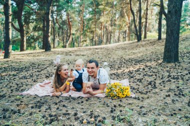 Genç ve çekici ebeveynler kırsal bir ormanda tatildeler. Şehir dışına gezileri çok seviyorlar. Küçük çocuklarıyla birlikte yatak örtüsünün üzerinde oturup kameraya bakıp gülümsüyorlar.
