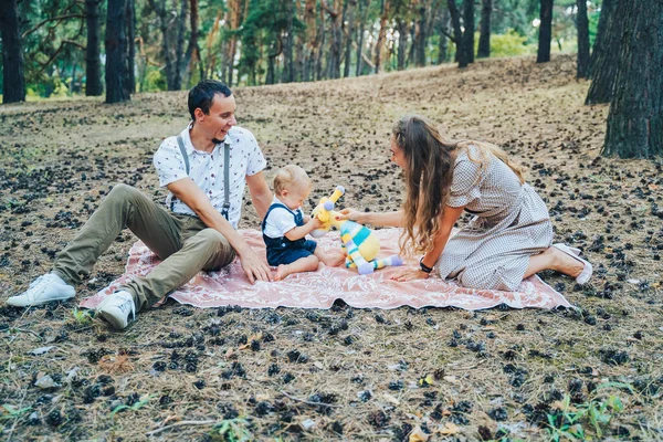 Happy family, wearing light clothes, sitting in a pine forest. Mom handing her son a soft toy of a giraffe. The kid is embarrassed. He likes the toy. Family entertainment concept. Time for each other