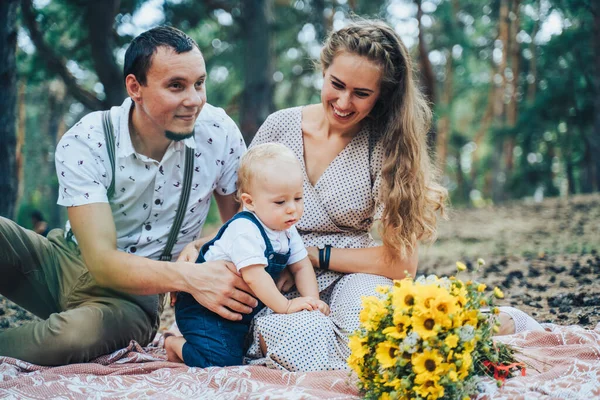 Beautiful family in beige clothes lies on a brown covering in a summer pine forest. Mom, dad and one year old baby. The kid examines, looking at flowers, cones, crawling on the ground