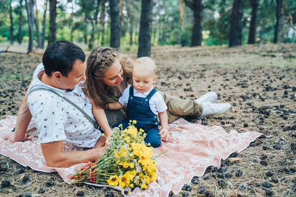 Beautiful family in beige clothes lies on a brown covering in a summer pine forest. Mom, dad and one year old baby. The kid examines, looking at flowers, cones, crawling on the ground
