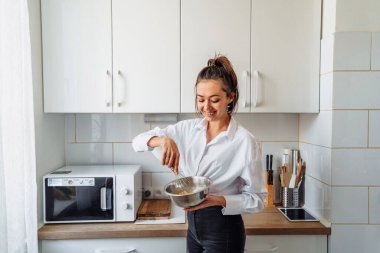 Güzel, çekici, gülümseyen, hamur yoğuran genç bir kadın. Çelik tabaklarda muz krepleri için çırpıyor. Beyaz, açık renkli bir mutfakta. Parlak aksansız tek renkli fotoğraf
