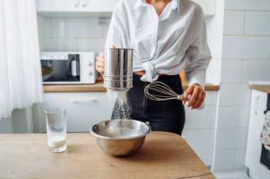 Ev yemeği konsepti. Parlak esmer bir kadın kekler için unu süzgeçten geçirir, hamuru tuzlar, süt ekler. Gülümseyen genç bir kadın, güzel, modern bir mutfakta yemek yapmayı seviyor..