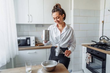 Ev yemeği konsepti. Parlak esmer bir kadın kekler için unu süzgeçten geçirir, hamuru tuzlar, süt ekler. Gülümseyen genç bir kadın, güzel, modern bir mutfakta yemek yapmayı seviyor..
