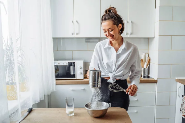 DIY home cooking concept. A bright brunette woman sifting the flour for pancakes through a sieve, salting the dough, adding milk. Young woman smiling, loves to cook in a beautiful modern kitchen.
