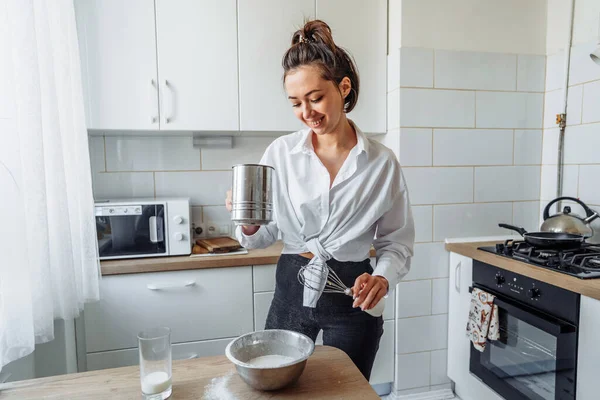 DIY home cooking concept. A bright brunette woman sifting the flour for pancakes through a sieve, salting the dough, adding milk. Young woman smiling, loves to cook in a beautiful modern kitchen.