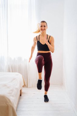 Zirvedeki güçlü atletik formalı ve tozlu kız, Enerjik bir şekilde koşuyor ve zıplıyor. Minimalist beyaz iç görünüşlü, Geniş ve parlak yatak odasındaki evinde gülümseyerek..