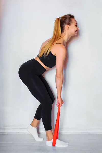 家庭でのスポーツトレーニングの概念 女の子はフィットネスガムを使用して腕や足のための演習を行います 強い抵抗力を持つ赤い色のゴムバンドで体内の筋肉を強化します — ストック写真
