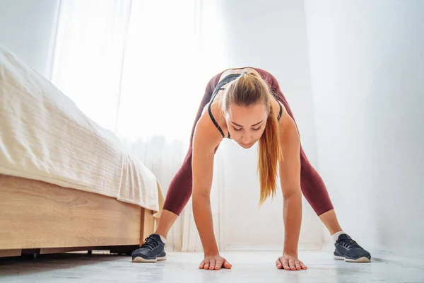 Beautiful pumped up girl is doing sports training at home. She tilting to different legs and leaning over down. Fitness workout at home in the bedroom near the bed in a bright, clear room