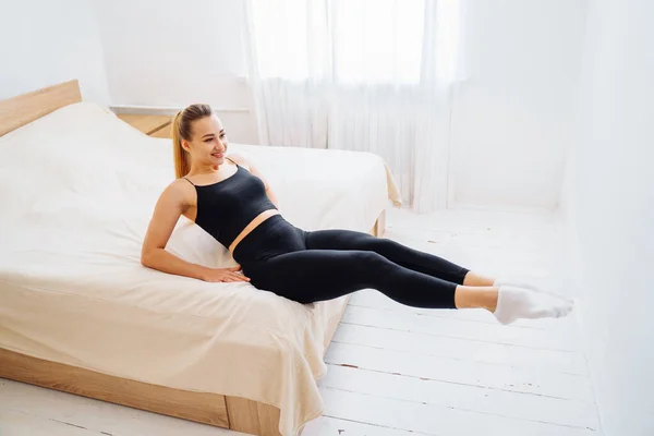 美しい運動女子 黒い服を着て 白いベッドの上に明るい光の色の寝室で自宅で室内プレスをポンピング 家に隔離されてる ジムのないフィットネス 健康的なスポーツの概念 — ストック写真