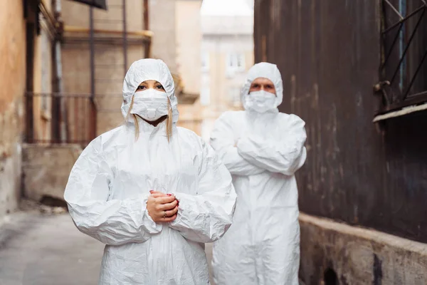 Doctors in protective suits and protective masks stand on the street with serious faces. Walking along the street, guy leads woman foward - they are very determined. The concept of coronirus covid-19