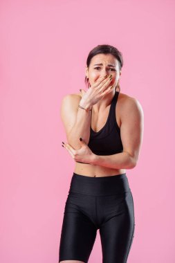 Siyah spor tişörtlü, yeşil gözlü esmer kadın. Şok olmuş yüzü, ağzını kapatıyor. Pembe duvardaki arka planda eli var.