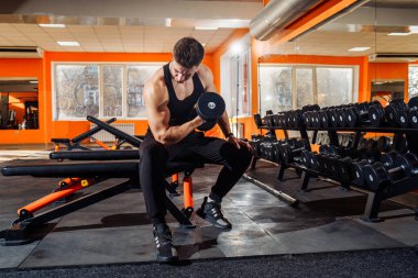 Atletik, yakışıklı, genç, kaslı Avrupalı esmer kızın portresi. Fiziksel egzersiz yapıyor, kol kasları spor salonunda, fotokopi odasında, halterlerle çalışıyor. Siyah tişört ve pantolon giyiyordu. Bakışları ve kasları