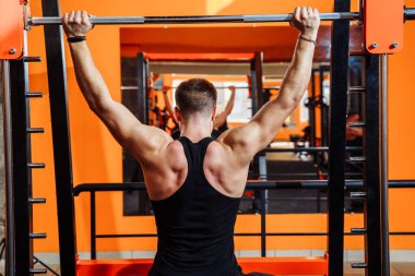 Ortalama plan, geriye bakış. Siyah spor kıyafetleri giymiş kaslı bir adam, çıtayı yükseltmek için kaslarını, pazılarını ve pazılarını çalıştırıyor. Spor ve spor. Sağlıklı yaşam tarzı kavramı