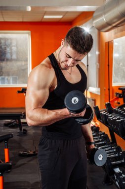 Spor salonunda ağırlık kaldıran güçlü, sağlıklı genç bir adam. Spor vücudu olan genç bir erkek spor salonunda egzersiz yapıyor. Dambılları kaldırıyor..