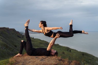 Açık havada tam boy portre genç çekici ve konsantre yoga akrobatları akroyoga yapıyor. Sporcu kadın erkek eli uzanıyor. Dünyanın kıyısında denge ve meditasyon egzersizleri.