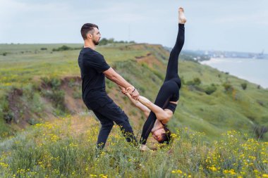Genç beyaz çift deniz kenarındaki tepede akro yoga yapıyor. Çift, partner yogası. Sporcu çok güzel esniyor ve eğitimli elleriyle kızı tutuyor. Sporcu kadın geriniyor