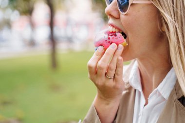Yüze yakın çekim, iştah açıcı bir şekilde pembe bir çöreği ısıran bir kadının dudakları. Parkta piknik yapan ve lezzetli bir tatlılığın tadını çıkaran kız. Ağzını kocaman açıyor.
