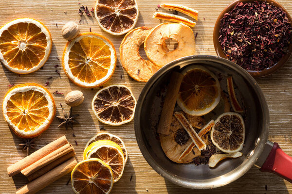 Top view of dry pieces of lemon, orange and apple, cinnamon sticks, bay leaves, nutmegs, star anise and dry cutted hibiscus as ingredients for tea and the saucepan