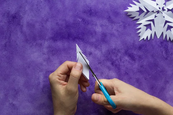 Woman hands cutting away shapes from the edges of the paper to shape snowflake pattern on the violet surface