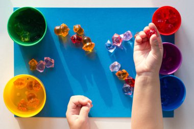 Çocuk eli rengarenk oyuncak taşlarıyla ayrılmış mavi yüzeye renkli bardaklara saçılmış.