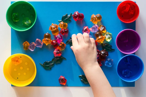 Mano infantil que separa por piedras de juguete multicolores de color dispersas en la superficie azul en copas coloridas —  Fotos de Stock