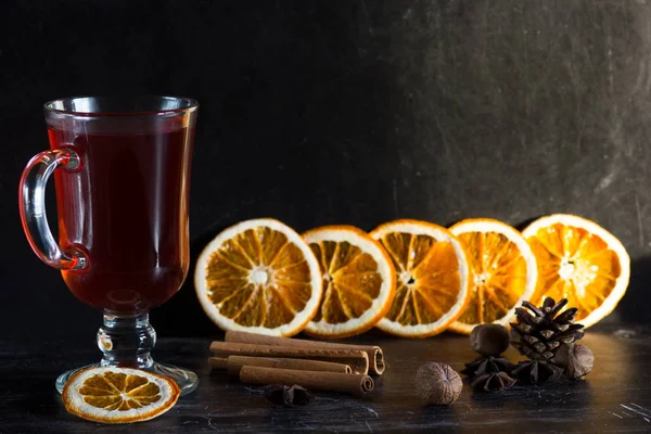 Copa de vidrio de vino caliente, especias y trozos de naranjas sobre el fondo negro — Foto de Stock