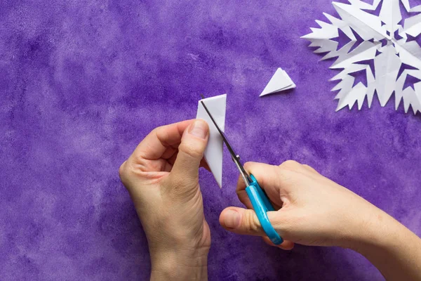 Woman hands cutting away shapes from the edges of the paper to shape snowflake pattern on the violet surface