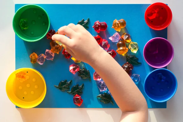 Дитяча рука розділяє кольорові різнокольорові іграшкові камені, розкидані на синій поверхні на різнокольорові чашки — стокове фото