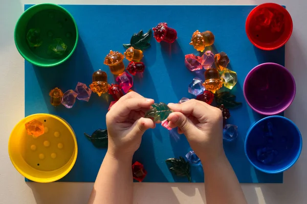 Child hand holding multicolored toys to separating into colorful cups by color on the blue surface — Stock Photo, Image