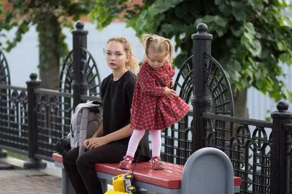 Mujer joven con mirada pensante sentada en el banco con bolsa con niño de dos años —  Fotos de Stock