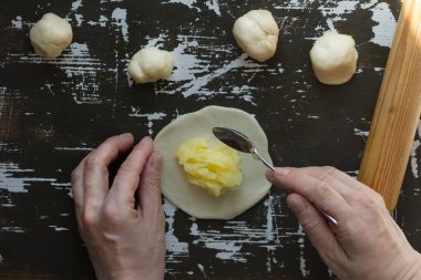 Kadın elleri yuvarlak hamurun üzerine patates püresi koyuyor. Kahverengi arka planda Vareniki için dolgu yapıyormuş gibi.