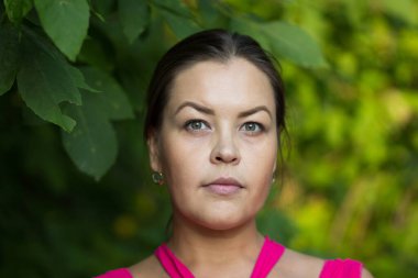 Yeşil yaz parkında gizemli ve düşünceli bir şekilde kameraya bakan orta yaşlı kadın portresi.