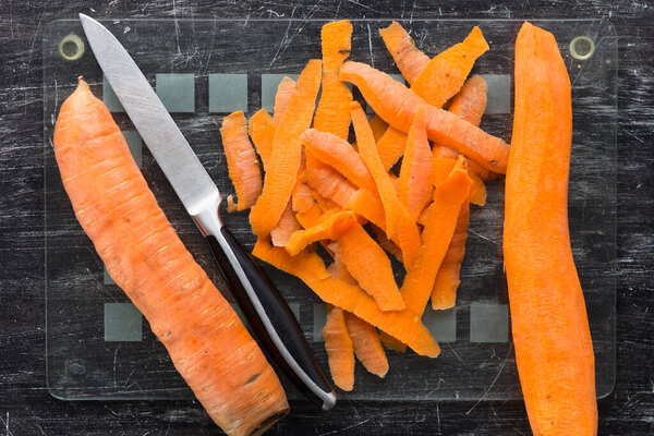 Top view of unpeeled carrot and peeled carrot with peeling and knife on glass cutting board on black background