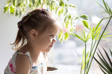 Üç yaşındaki üzgün beyaz çocuk pencere eşiğinde oturup pencereye bakıyor.