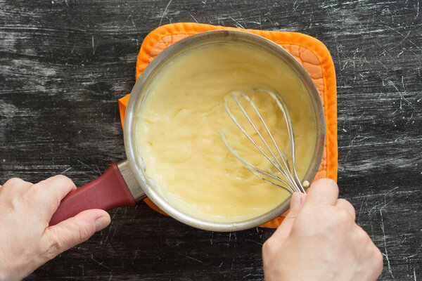 Top view of woman hands stirring hot thick mix of yolks, starch, sugar and milk making custard cream on black background