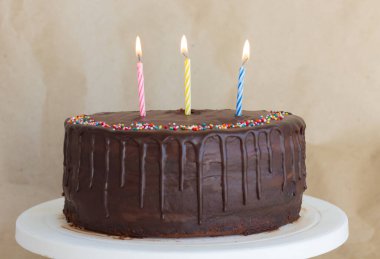 Üç mum ve beyaz tabakta çikolata damlalarıyla çikolatalı pasta.