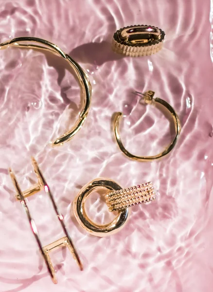 Arany karkötők, fülbevalók, gyűrűk, ékszerek rózsaszín víz backgr — Stock Fotó