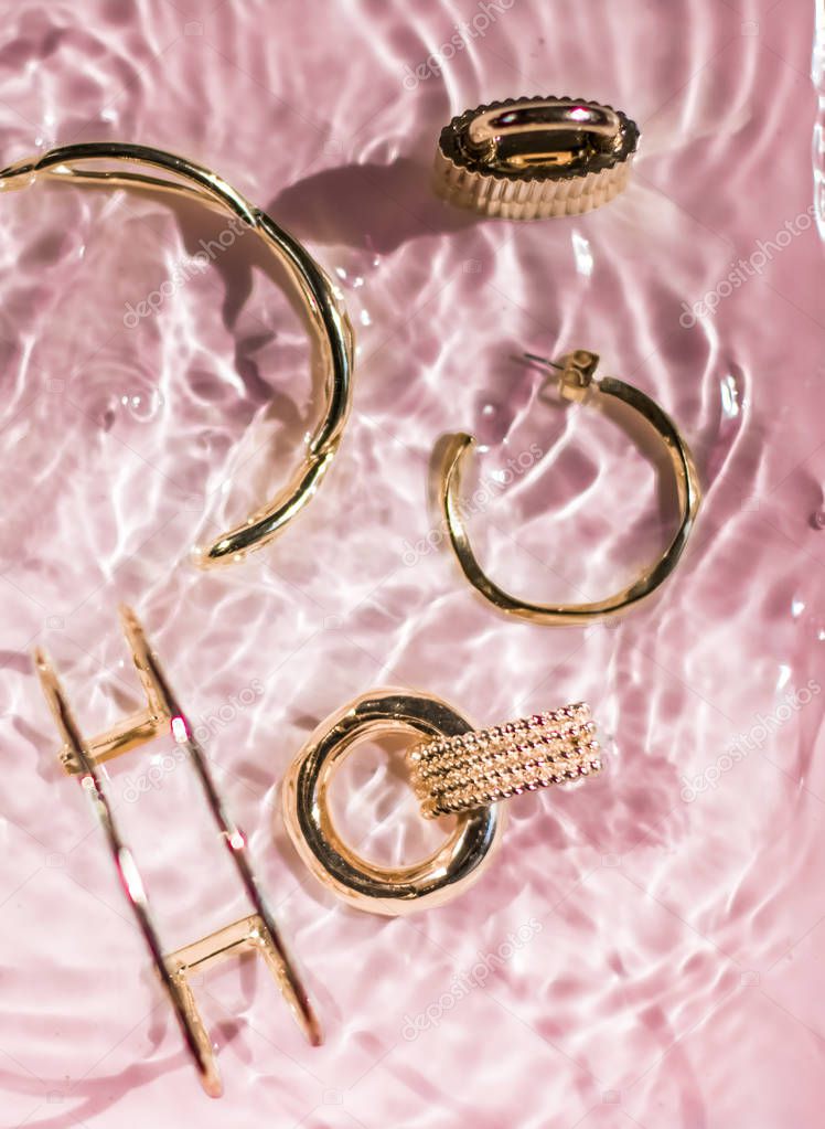 Golden bracelets, earrings, rings, jewelery on pink water backgr