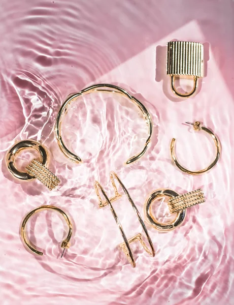 Golden bracelets, earrings, rings, jewelery on pink water backgr
