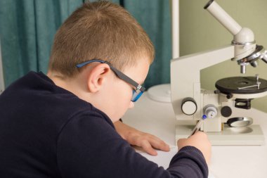 Gözlüklü ve ceketli çocuk masaya oturur ve okul ödevini yapar.