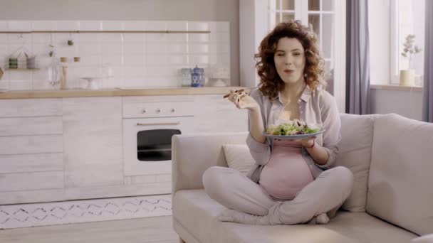 妊娠中の女性は台所でソファに座りながらサラダを食べる — ストック動画