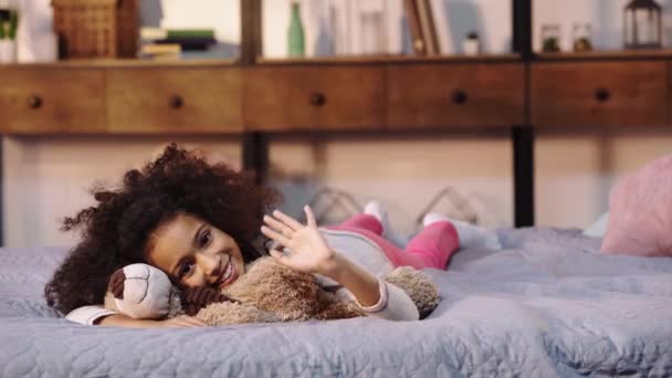Χαρούμενο Αφροαμερικάνικο Παιδί Κουνάει Χέρι Ενώ Ξαπλώνει Αρκουδάκι Στο Κρεβάτι — Αρχείο Βίντεο